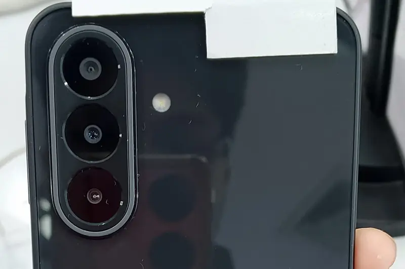 Back of a black Samsung Galaxy A27 5G held in a hand in a store, showing its triple camera, with a blank white tag taped across the top