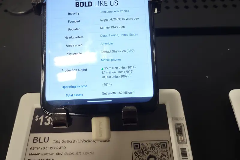 A BLU G64 phone clamped to a display at a Best Buy store in New York, above its electronic shelf label with a dollar price starting "$13"