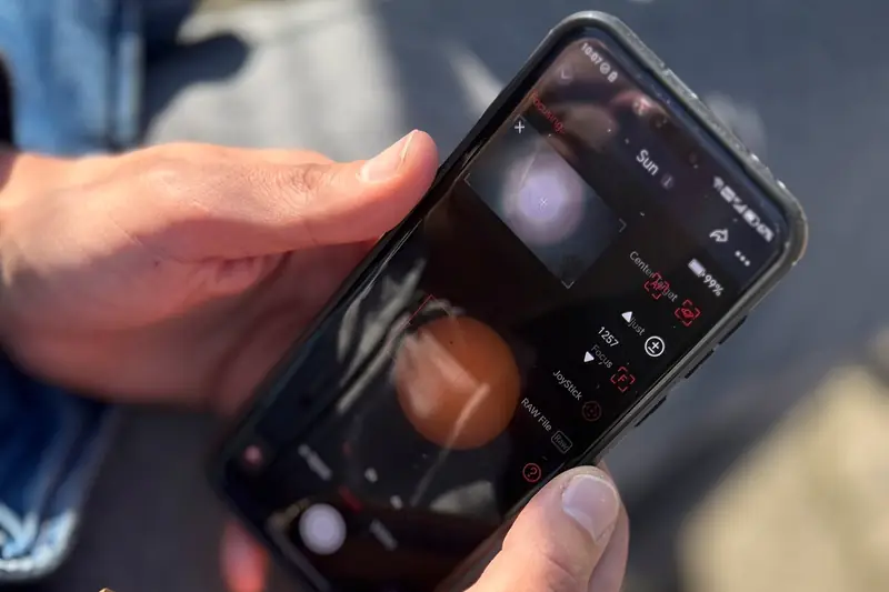 Two hands hold a smartphone in bright sunlight, with a denim jacket and a bag in the background
