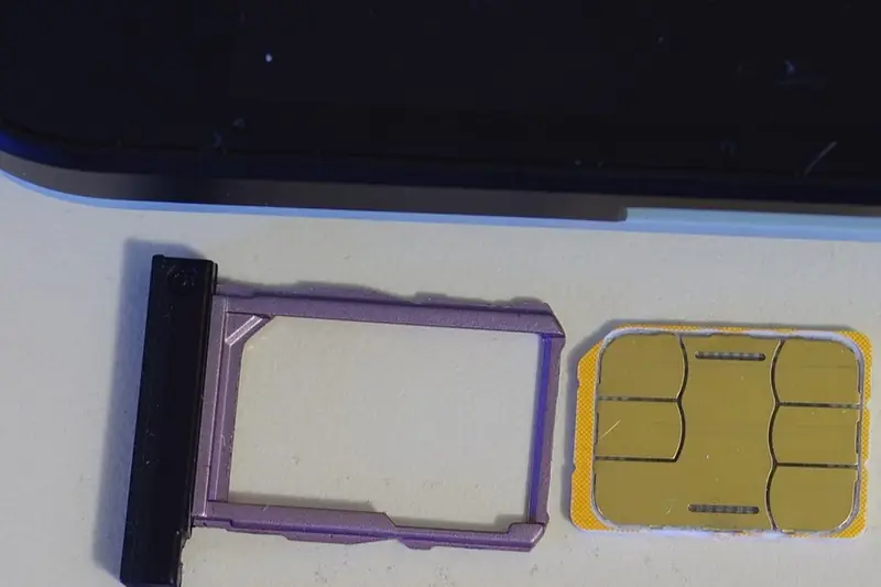 A yellow nano-SIM card lying beside an empty purple SIM tray under the bottom edge of a dark smartphone